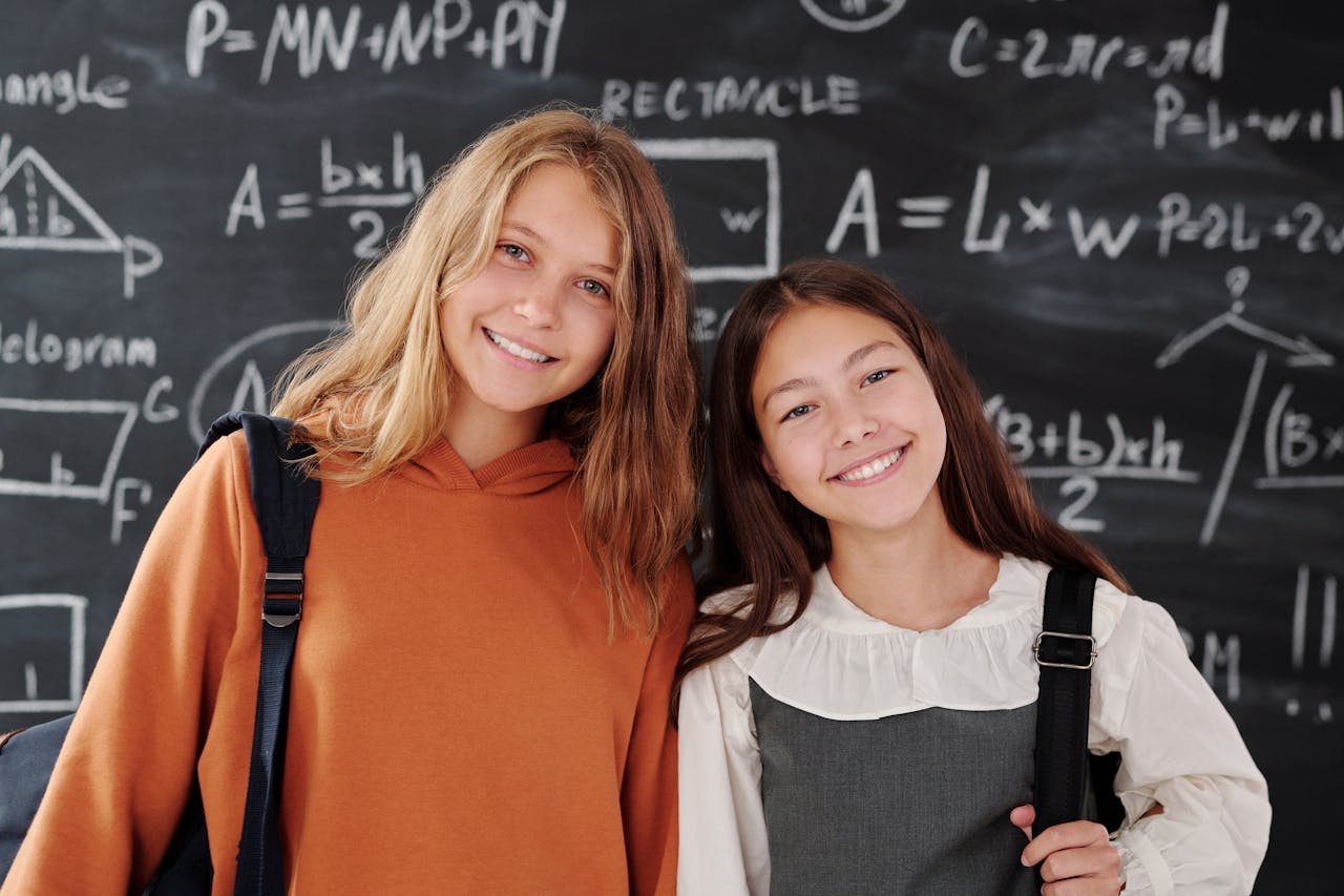 Home Two cheerful girls stand in a classroom with a math-covered chalkboard, ready for learning.